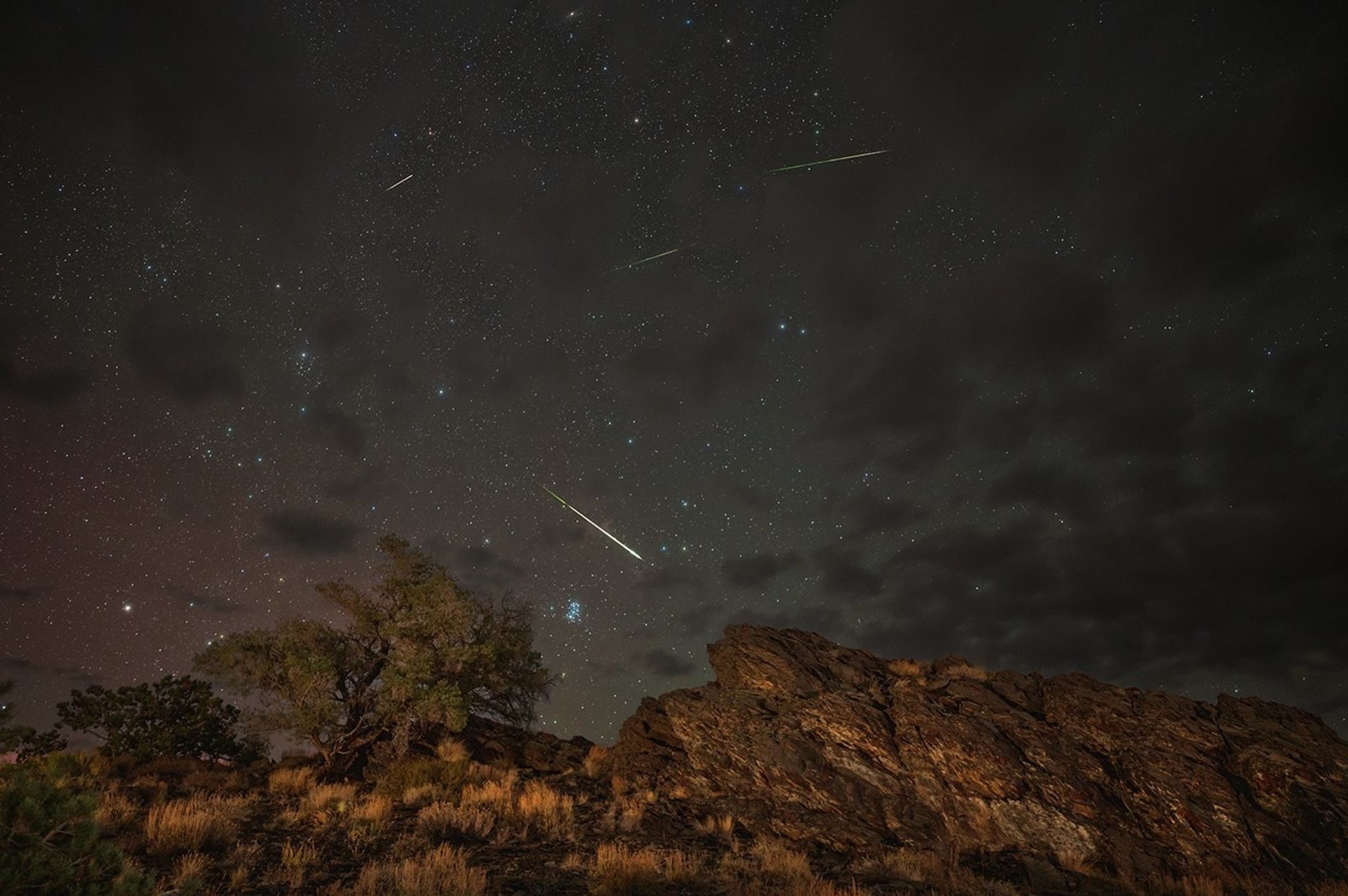 Four yellowish meteors streak across a partly cloudy sky above some green trees and brown rocky, hills.