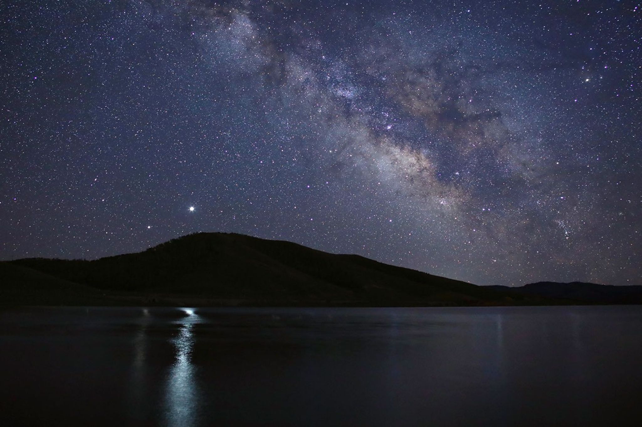 Saturn and Jupiter appear as two bright stars above a mountain ridge, reflecting brightly on the surface of a lake