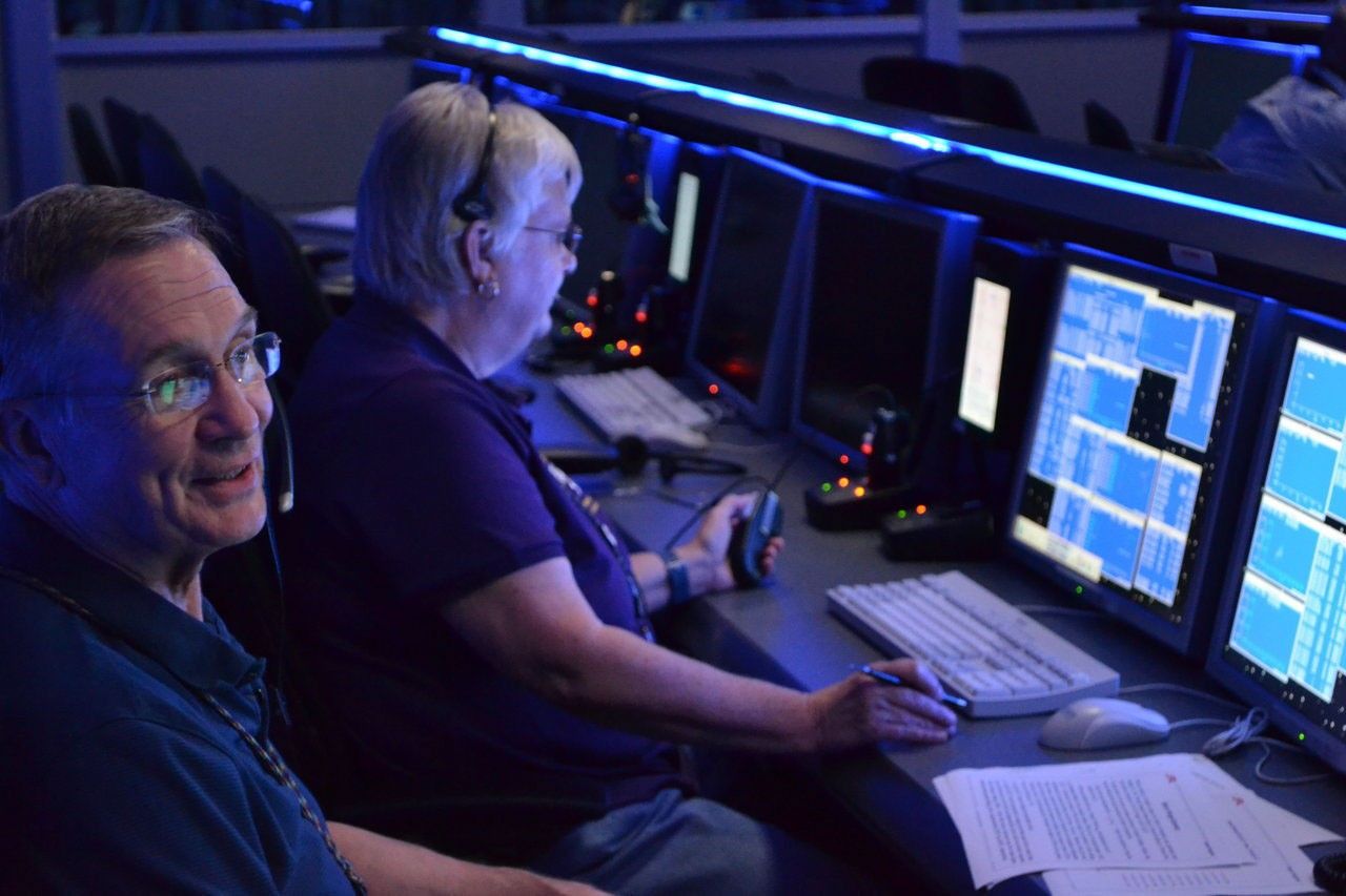 Color image of man and woman sitting at a bank of computer consoles.