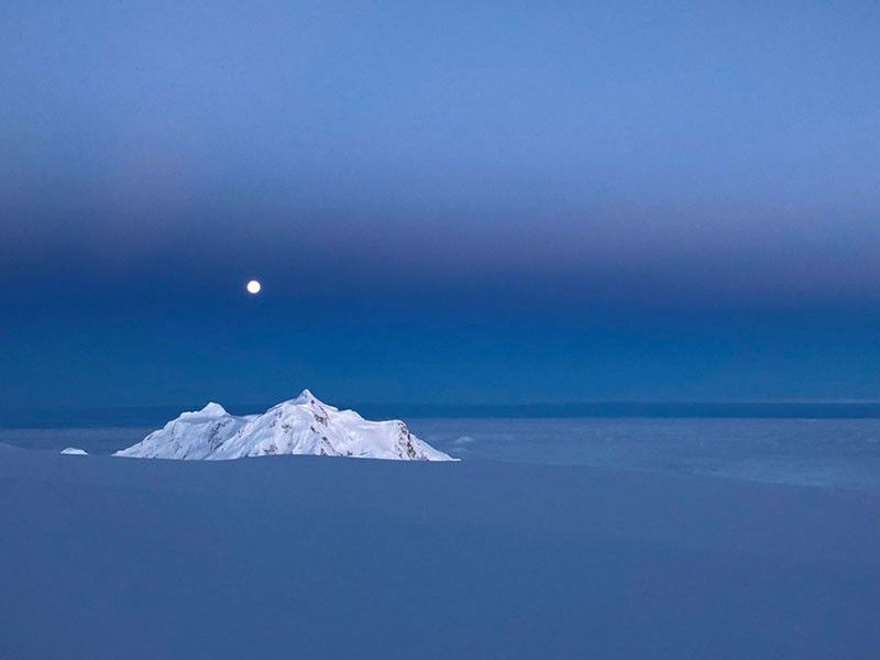 La luna llena se eleva sobre un pico nevado en Alaska.