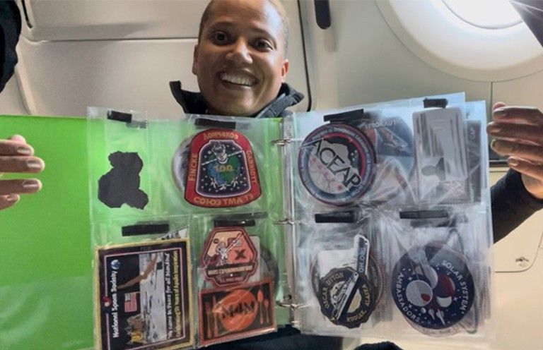 A smiling Dr. Sean Proctor floats inside a spacecraft with a notebook floating in front of other that is opened to show clear plastic pages with pockets holding badges and patches.