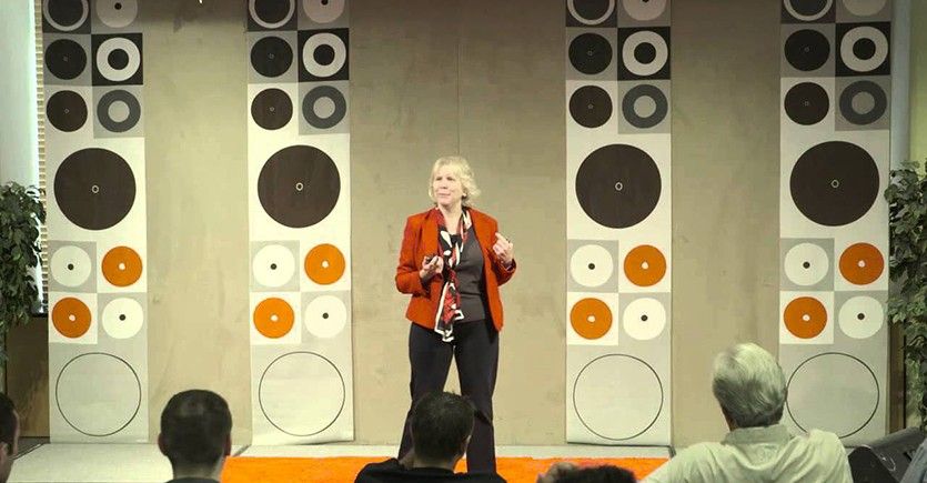 Darby Dyar wears an orange blazer, dark slacks, and a multi-colored scarf as she stands on a stage with decorative panels behind her that have circles of various sizes. The backs of heads of some spectators in the audience also can be seen.