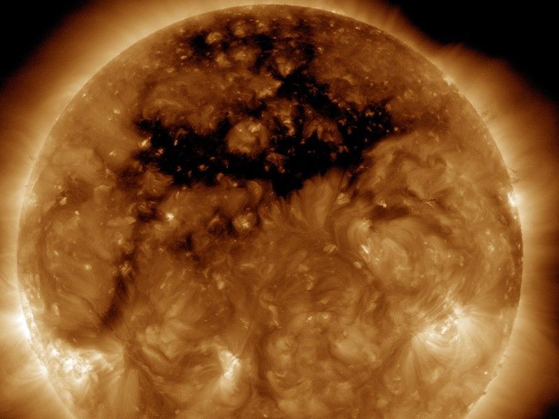 The Sun appears in patchy shades of gold with some brighter and darker regions, set against a black background. In the upper part of the Sun is a large, irregularly shaped dark area known as a coronal hole.
