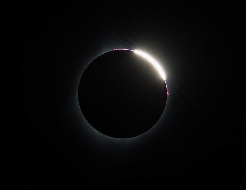 The eclipsed Sun, appearing as a black circle with a faint white glow. On the upper area, bright white and pink areas peak out from behind the Moon.