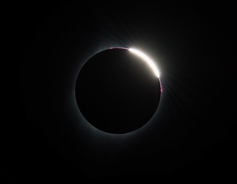The eclipsed Sun, appearing as a black circle with a faint white glow. On the upper area, bright white and pink areas peak out from behind the Moon.