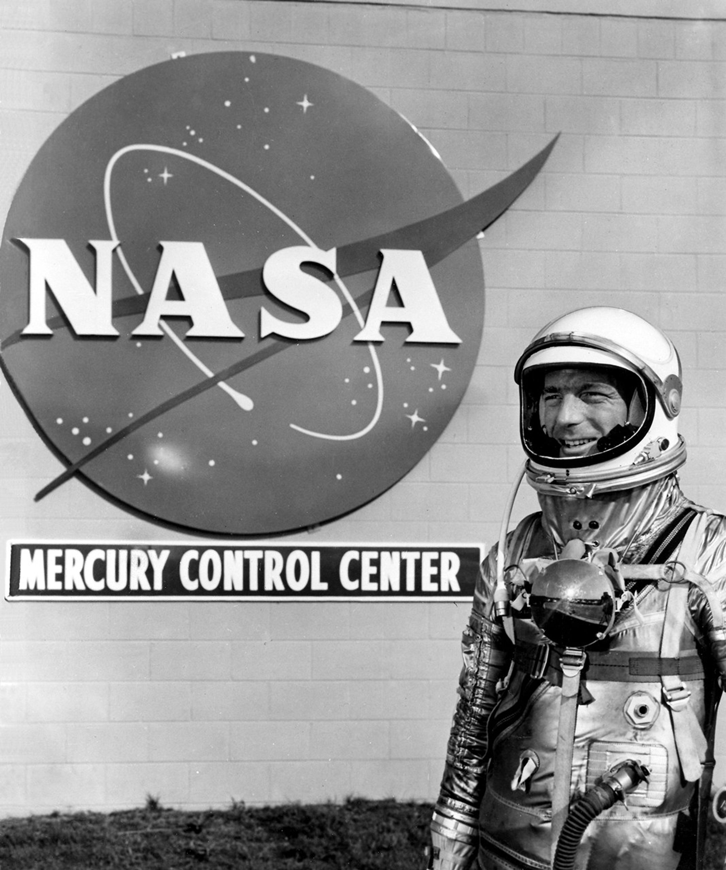 A black and white image of an astronaut stands in front of a building that holds a large NASA Meatball logo and a sign reading, "Mercury Control Center."