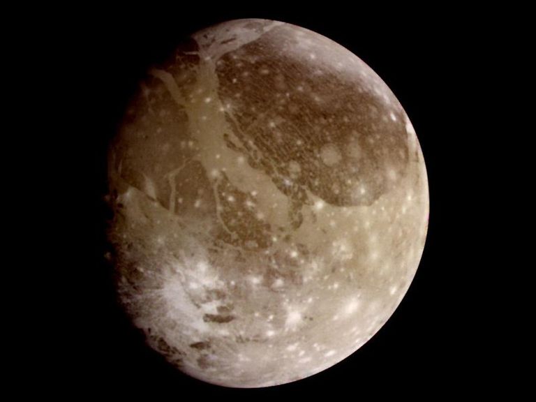 A brownsh gray sphere, shaded on the left side, appears against a black background. It's mottled with dark patches in the upper half, while the lower half is spotted with beight, white mars, some with rays extending from them like paint splatters.