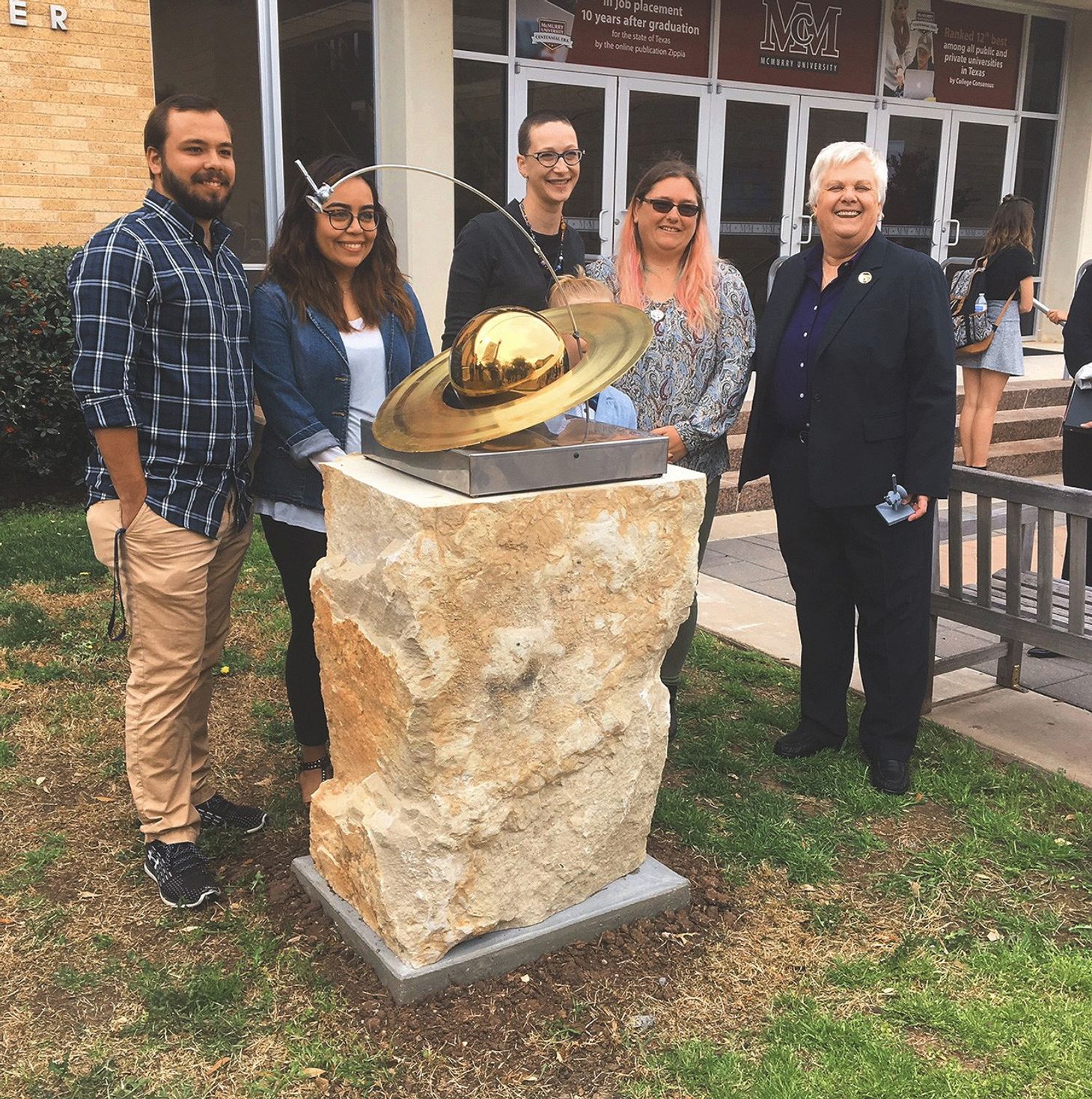 Group of smiling people around statue of spacecraft and Saturn.