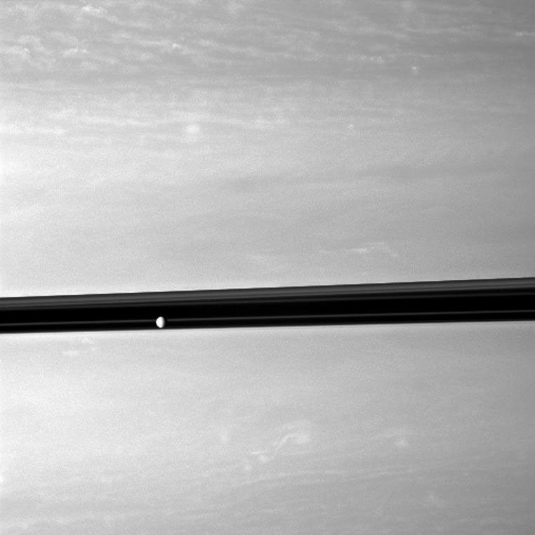 Saturn's moon Enceladus orbits serenely before a backdrop of clouds roiling the atmosphere the planet.