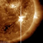 A portion of the Sun is shown in shades of gold. A large white flash of light, a solar flare, appears on the Sun at the center of the image.
