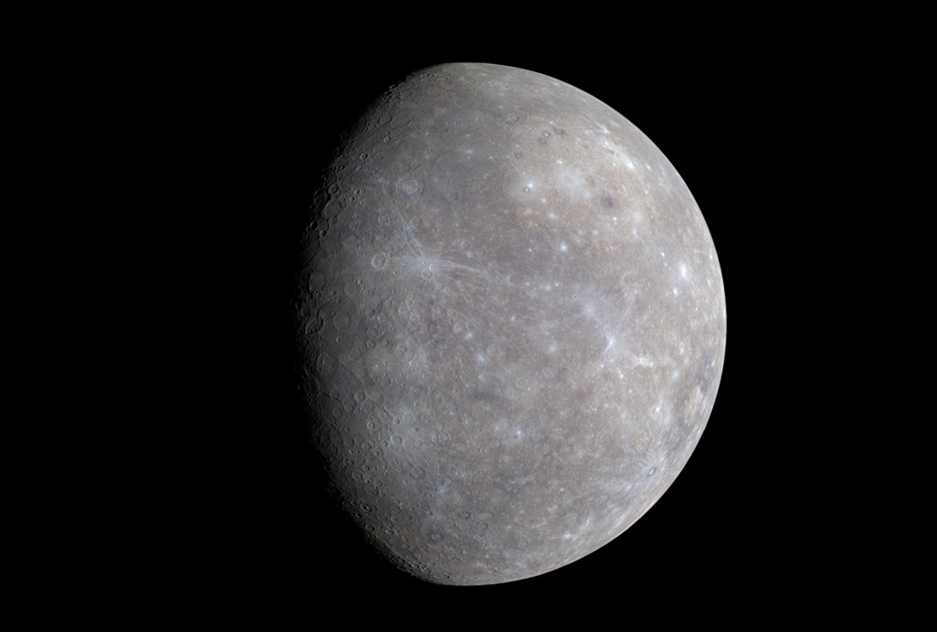 A gray, cratered world in the blackness of space, with about two-thirds of the planet illuminated by sunlight coming from the right. The overall gray shows some tan tints in some areas, and several craters have slivery-white rays extending outward from them, across the surface.