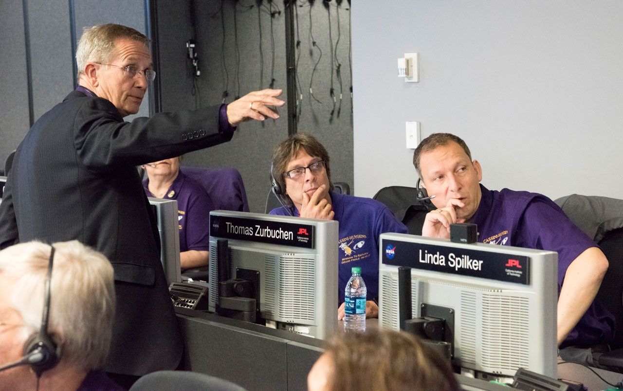Three men in mission control looking at data on a big computer screen.