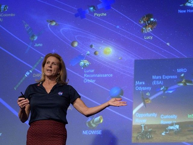 A woman stands in front of a large screen, gesturing at an image with her left hand.