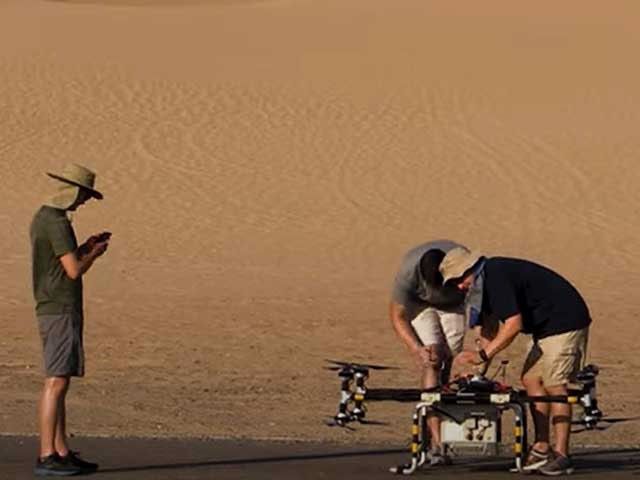 Two humans are bending over a model spacecraft. A third human is shown watching at the left. A sandy surface is shown in the background.