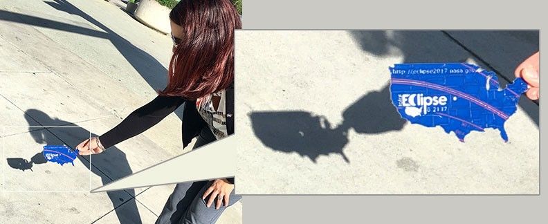 A woman holds a pinhole projector in her hand. The project is shaped like the U.S. and has a small hole in the center. Behind the projector on the ground, there is the shadow of the projector. In the middle, a small, crescent of light appears. This is the shape of the Sun.