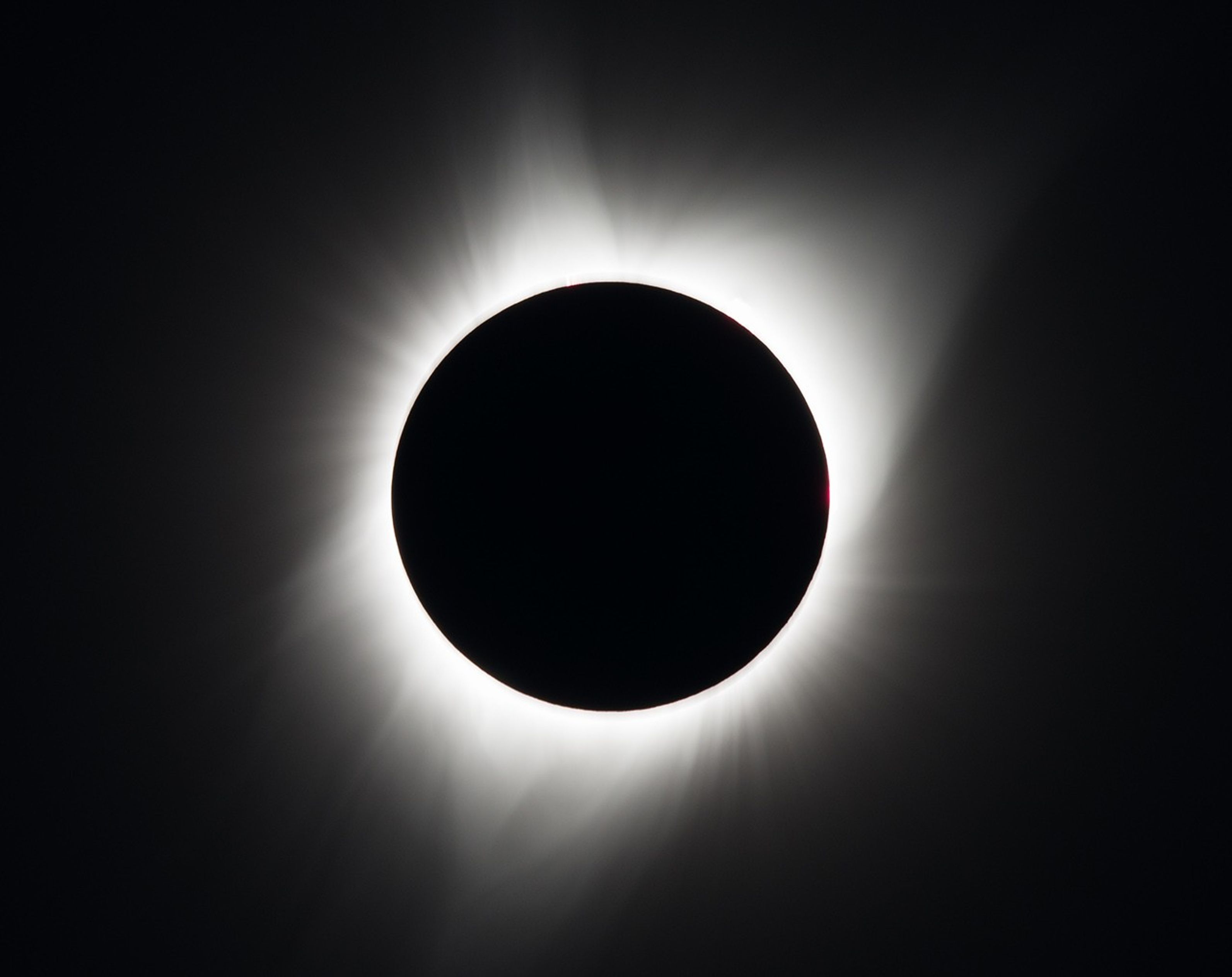 The Moon appears as a black disk in the center, surrounded by a white, asymmetrical halo of wisps and streamers, set against a black background.
