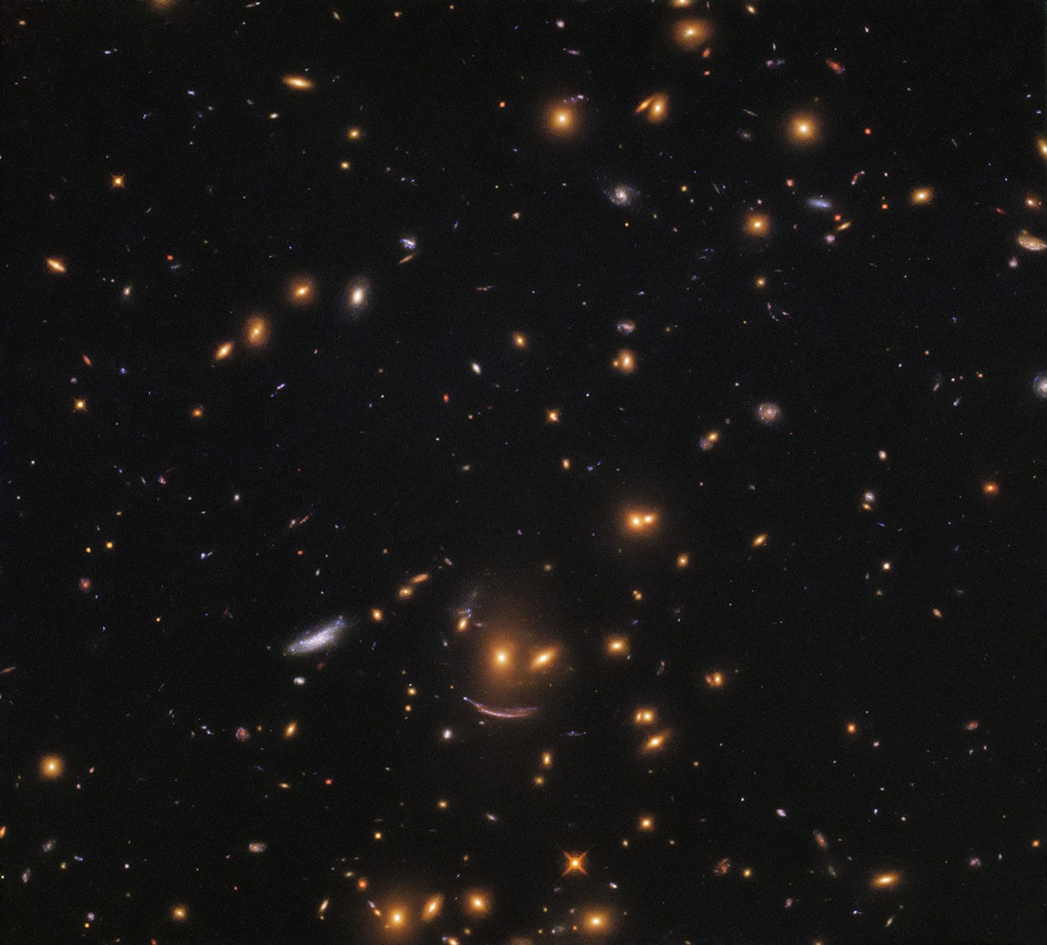 Three galaxies look like a smiling fact with two yellow dots for eyes and a stretched (gravitationally lensed) galaxy makes up the smile.